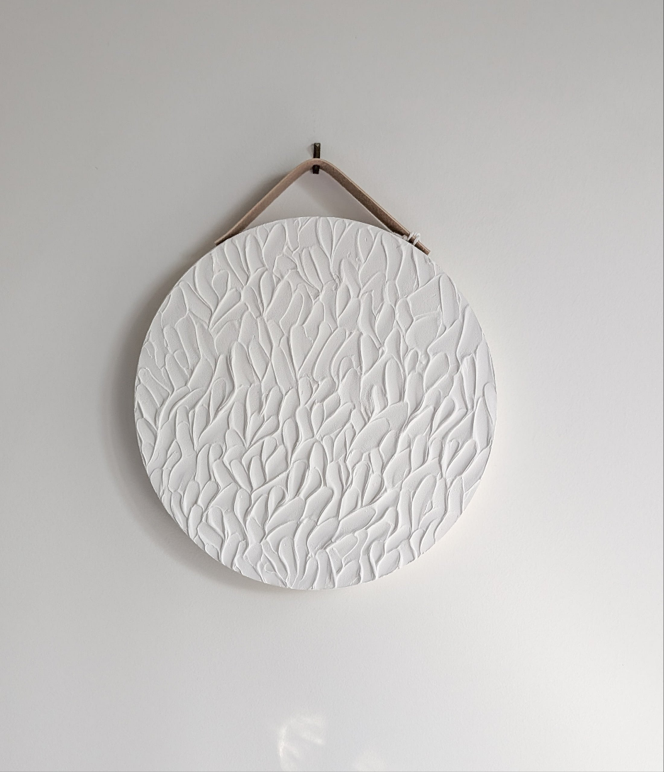 Close-up of white circular textured artwork with flowing leaf stroke patterns, suspended from a tan strap on a clean neutral wall – serene handmade wall decor by Christen Rose.