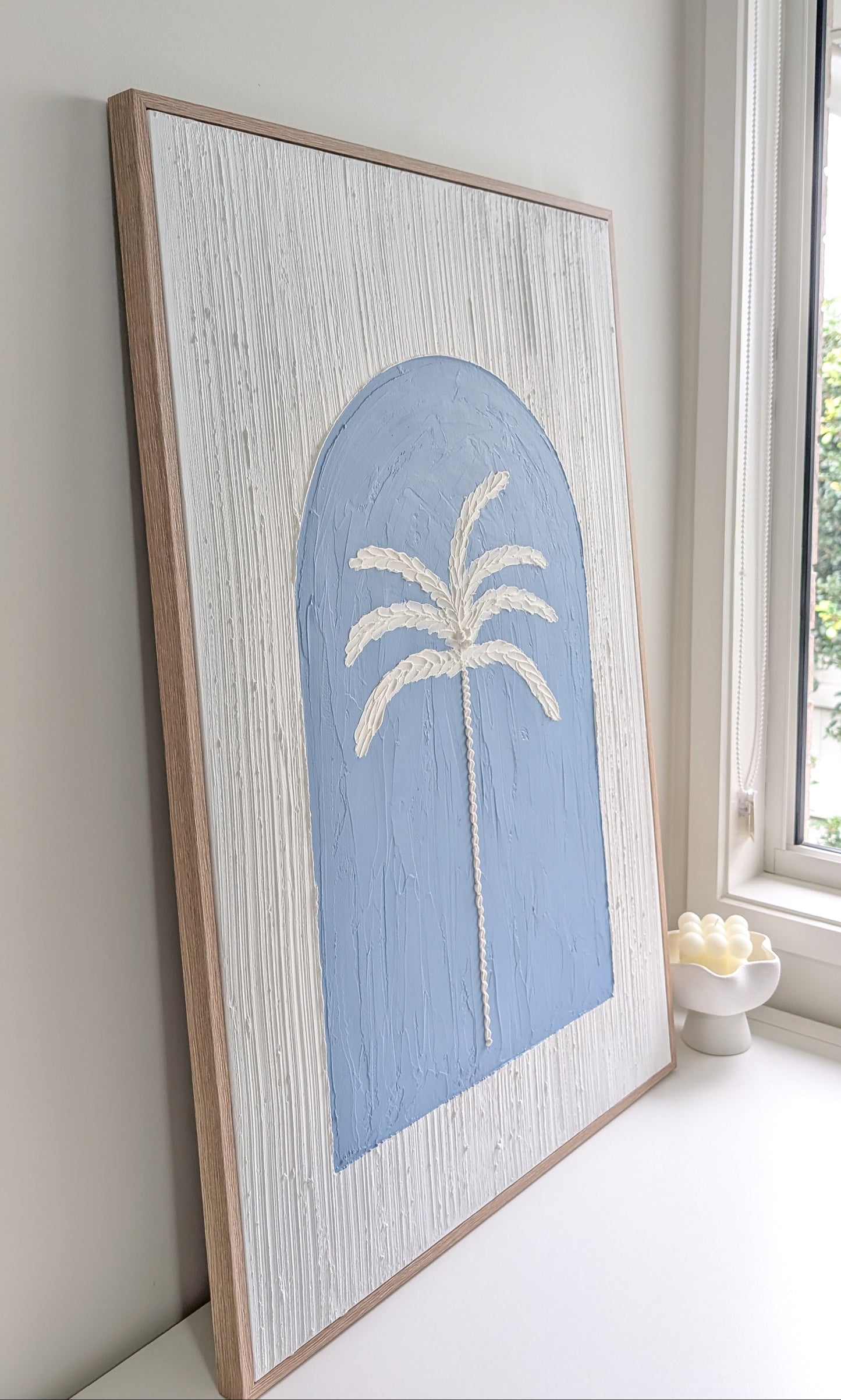 Framed artwork of a palm tree with a blue arch on a white wall.
