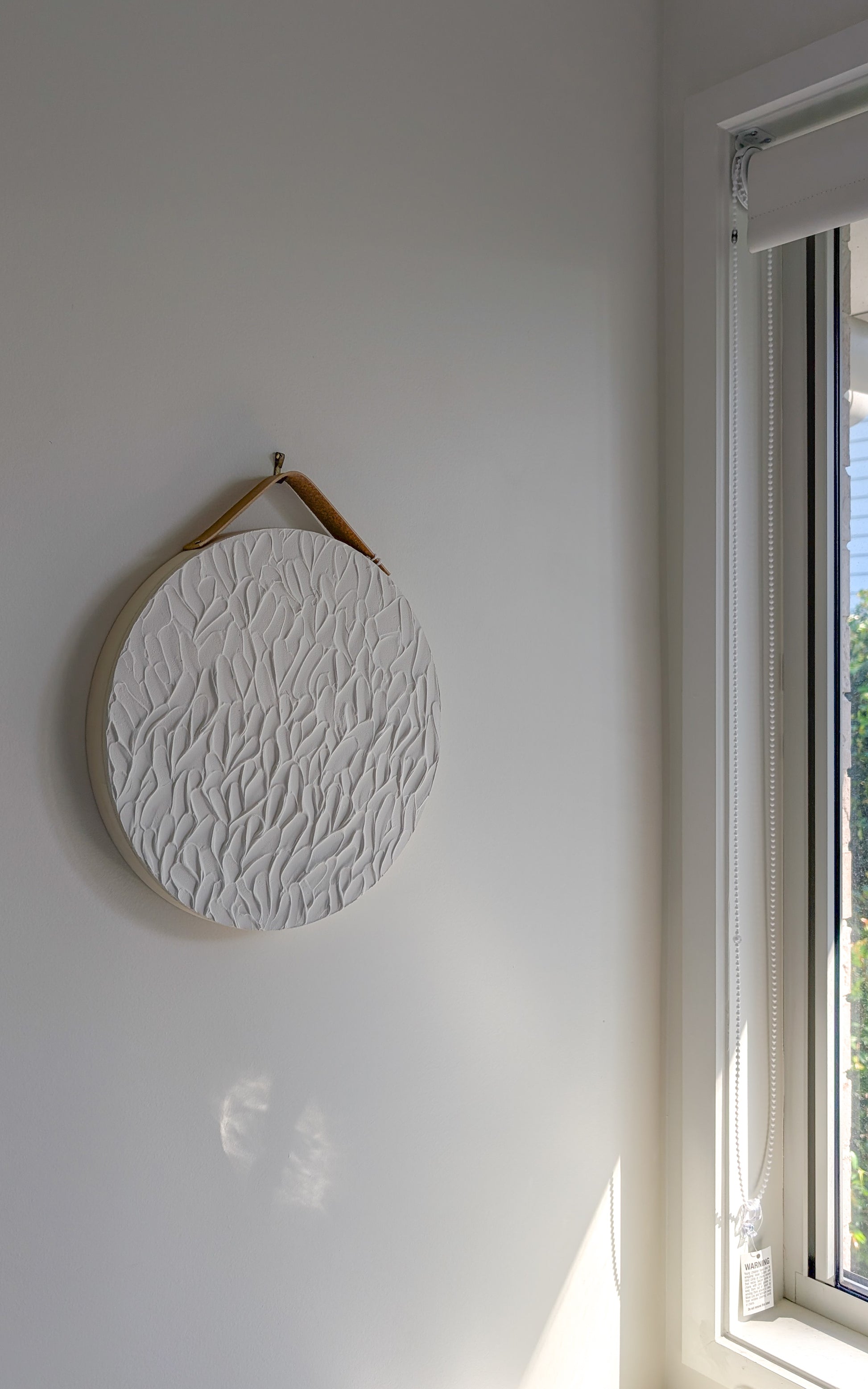Round textured wall art on a white wall near a sunlit window, featuring raised leaf-like strokes and a faux leather hanging strap – coastal minimalist design by Christen Rose.