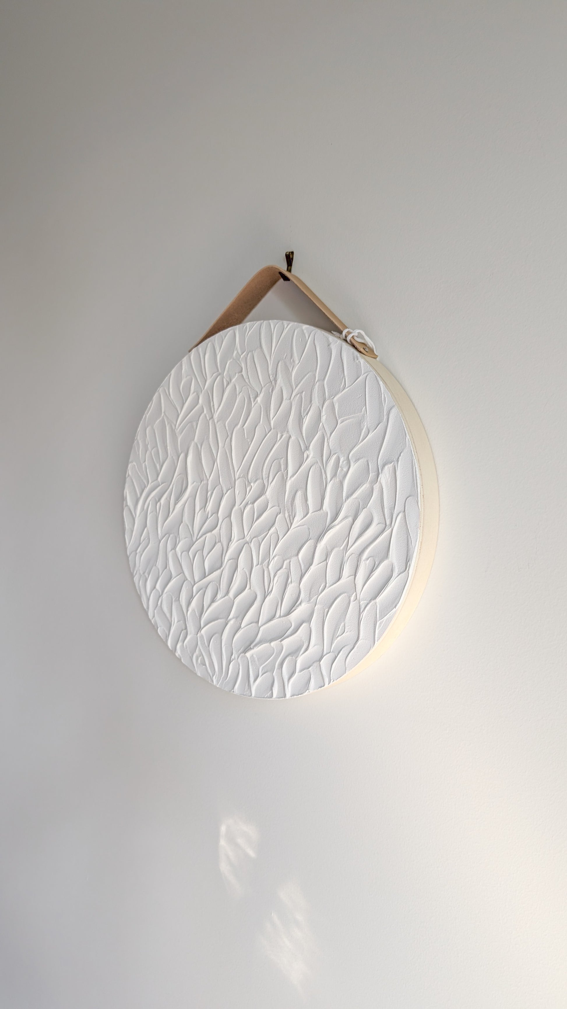 pure white textured art leaf stroke design on a round wood panel hanging on a light grey wall background