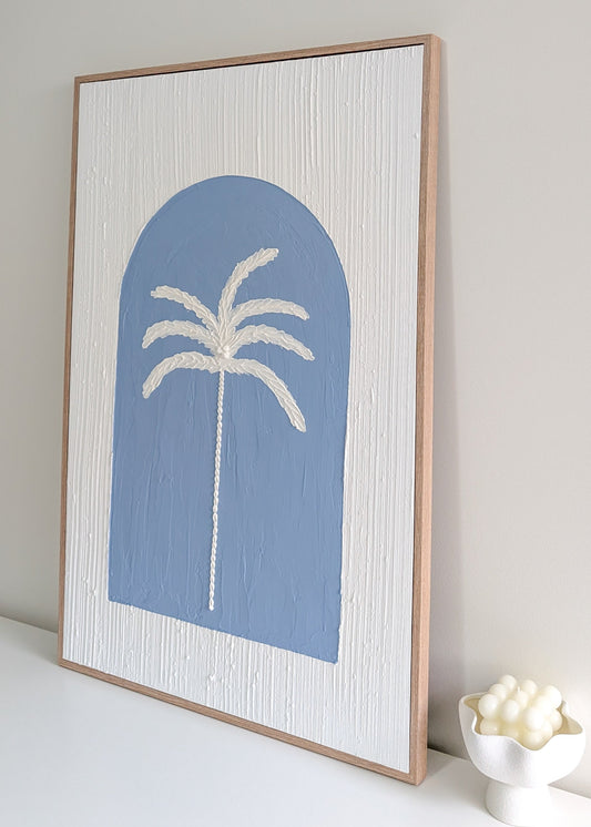 Framed artwork with a white palm tree inside a blue arch design on a light gray wall.