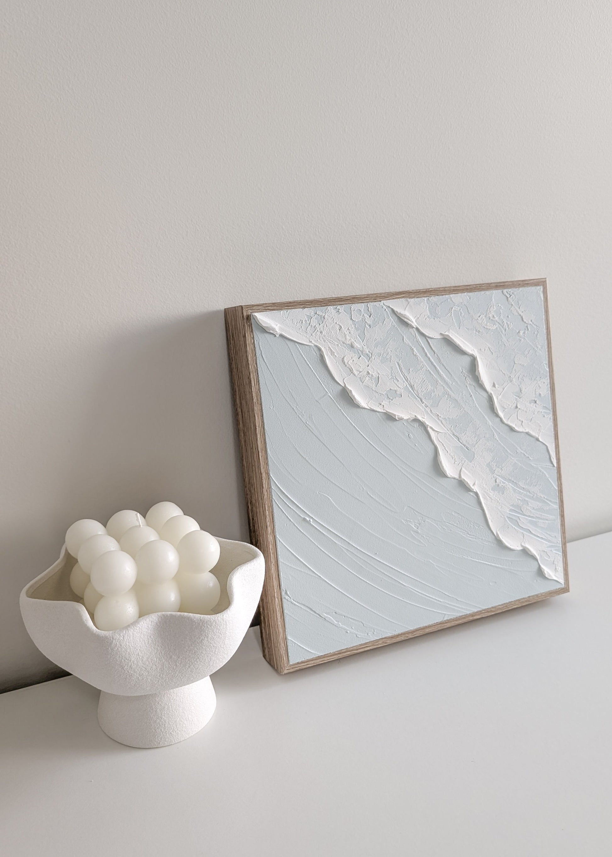 Framed coastal ocean inspired artwork on a wall next to a white bowl and white candle