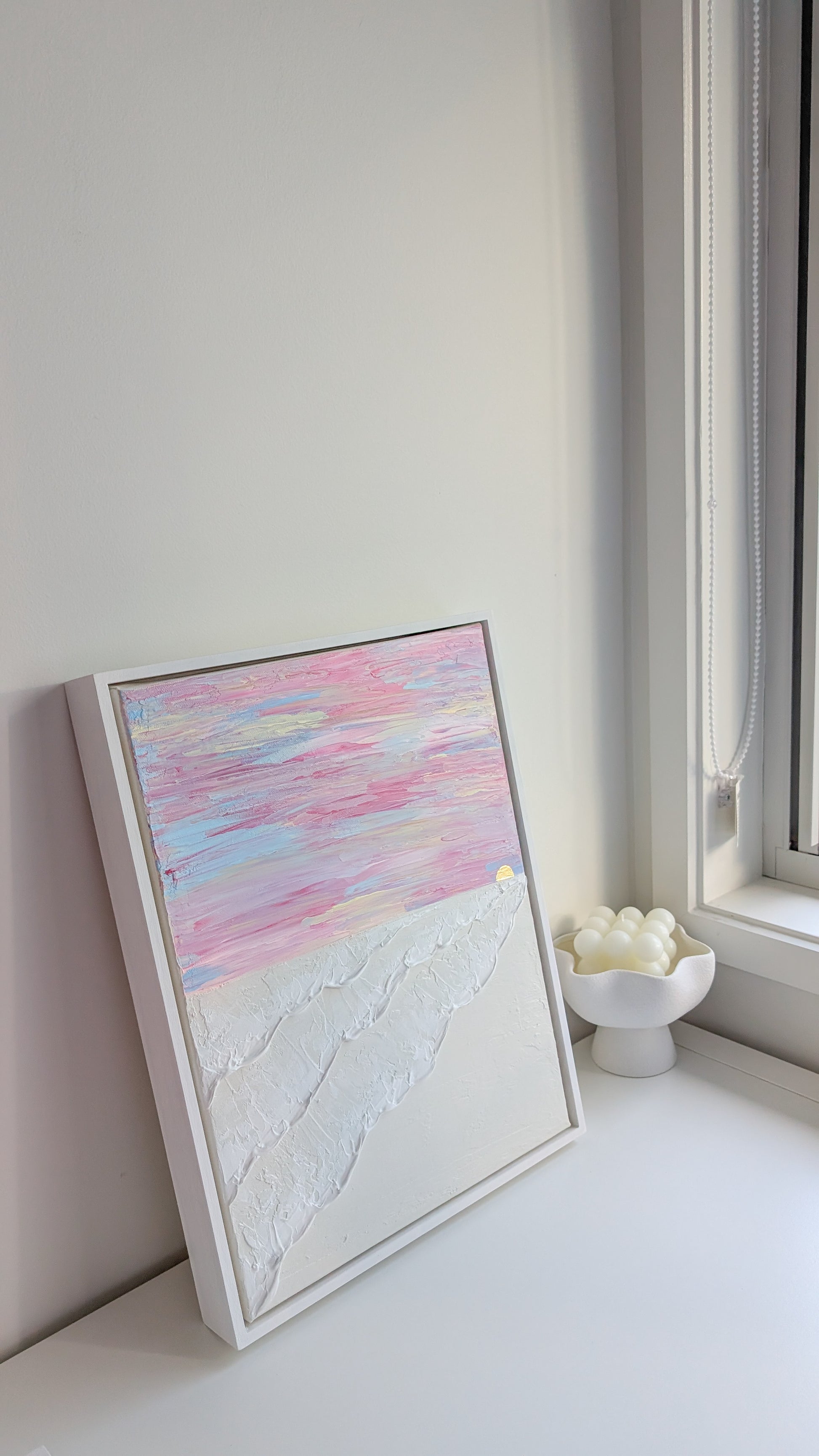 Handmade textured painting of ocean waves and a pastel sunset, leaning against a window with coastal decor.