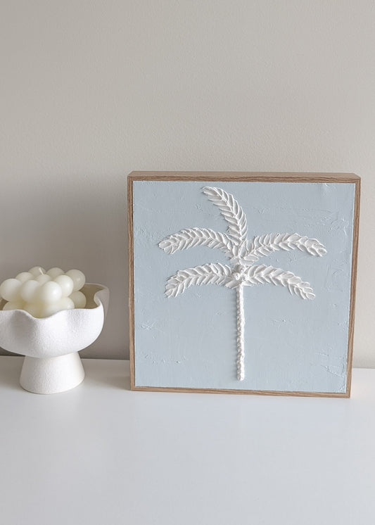Blue palm tree textured wall art in wood-look frame, styled on neutral wall with seashell decor