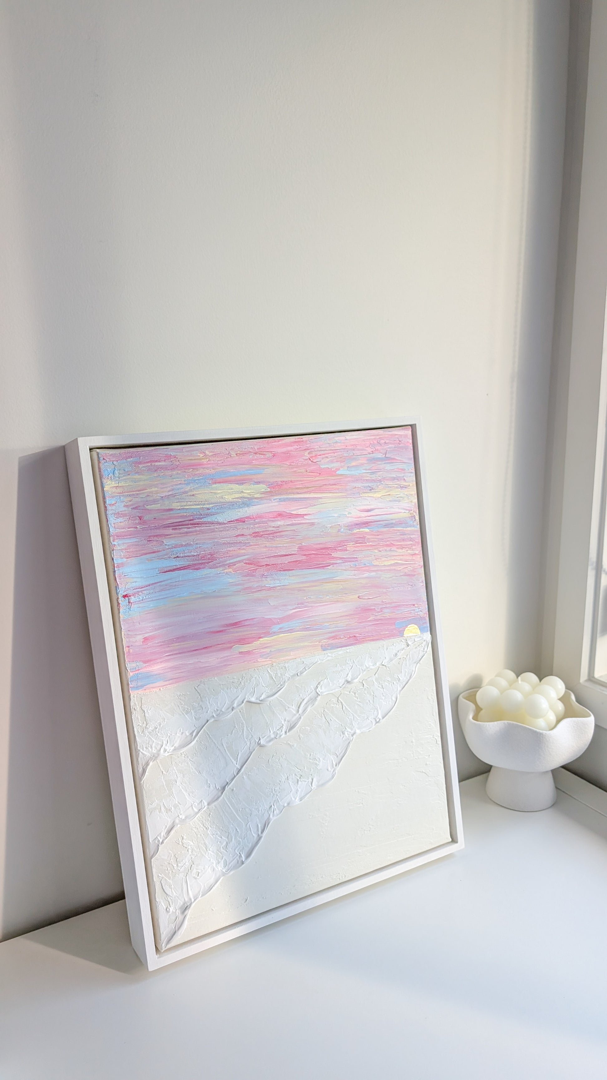 Textured coastal wall art in pink and white pastel tones, framed in white wash wood, displayed on a white shelf.
