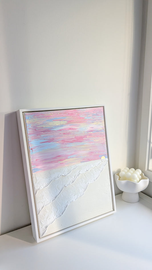 Textured coastal wall art in pink and white pastel tones, framed in white wash wood, displayed on a white shelf.
