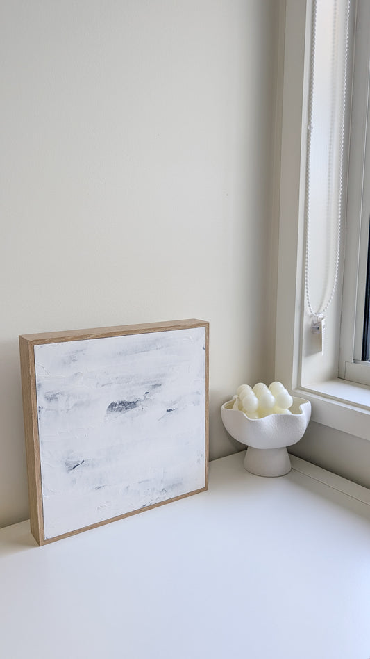 Minimalist textured abstract artwork in blue and white tones, framed in natural wood, styled on a windowsill.