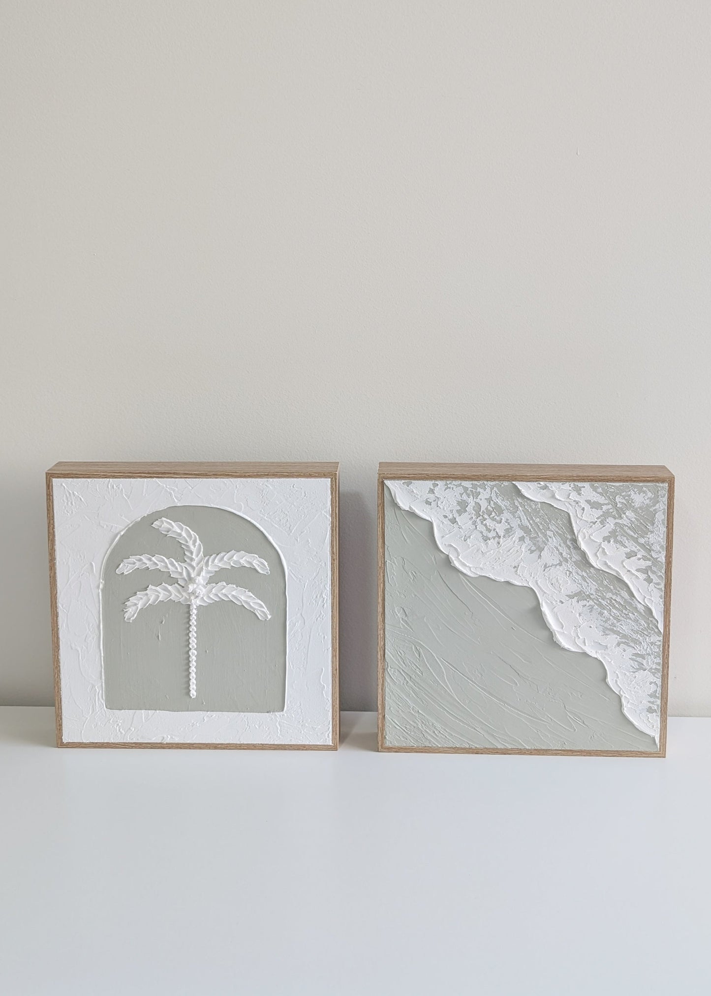 Two framed prints of a palm tree and beach scene on a white surface with a light gray wall background.