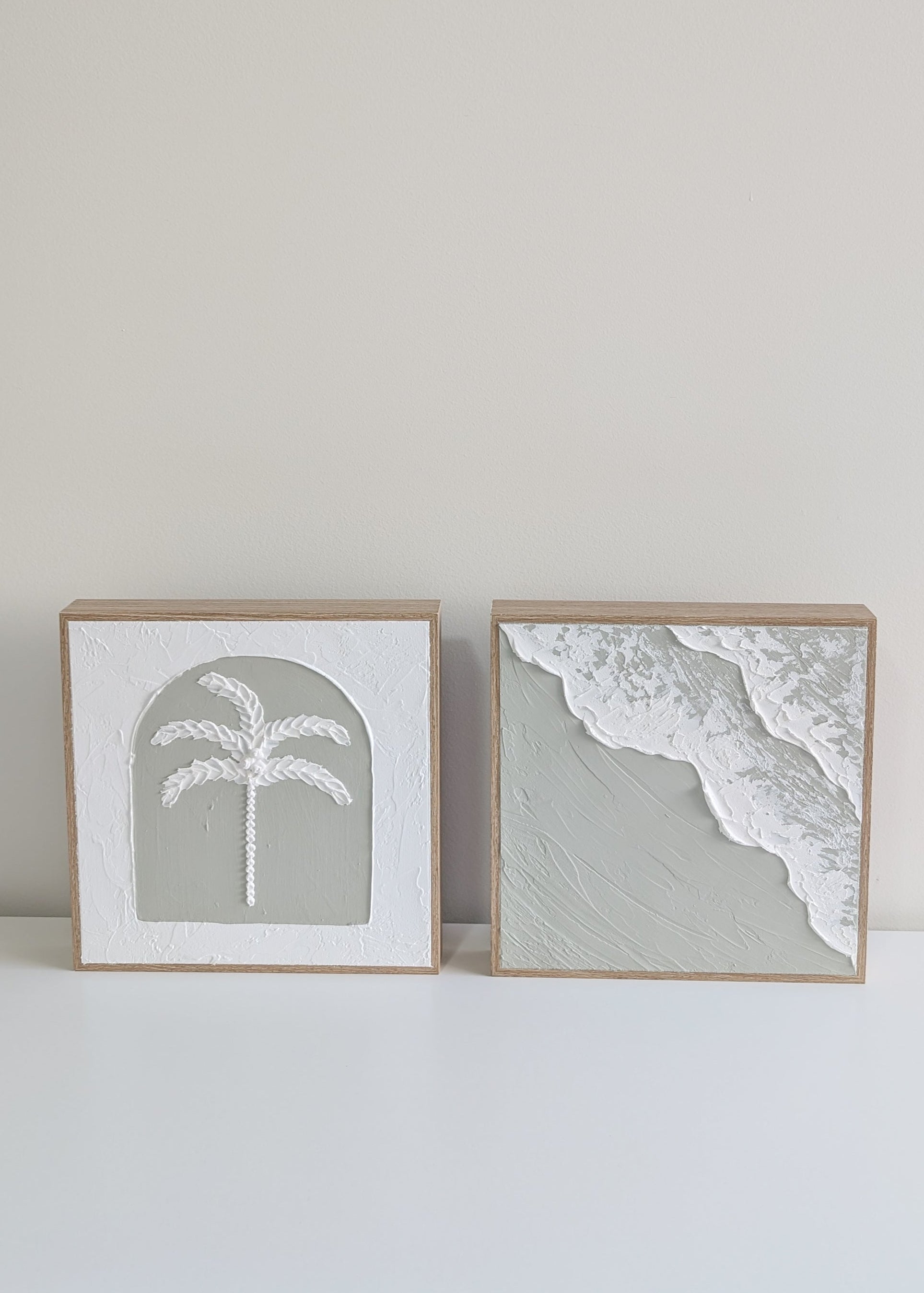 Two framed prints of a palm tree and beach scene on a white surface with a light gray wall background.