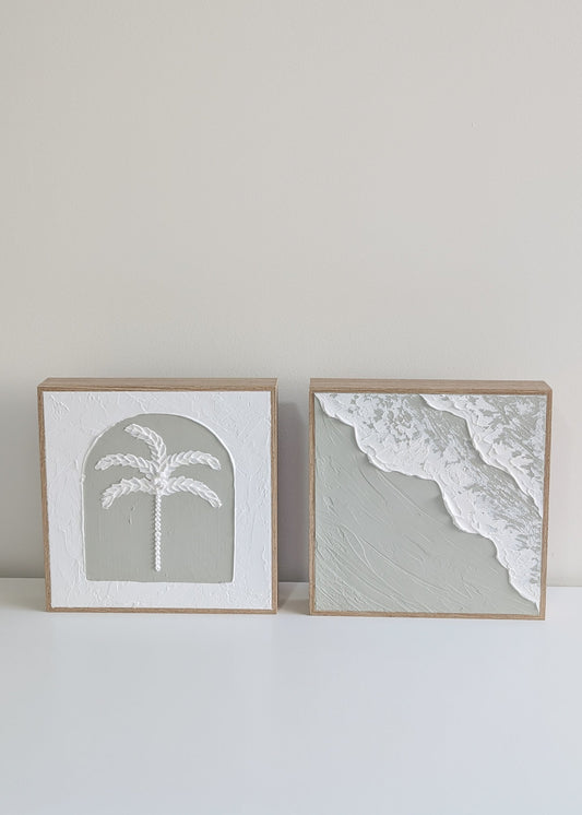 Two framed prints of a palm tree and beach scene on a white surface with a light gray wall background.