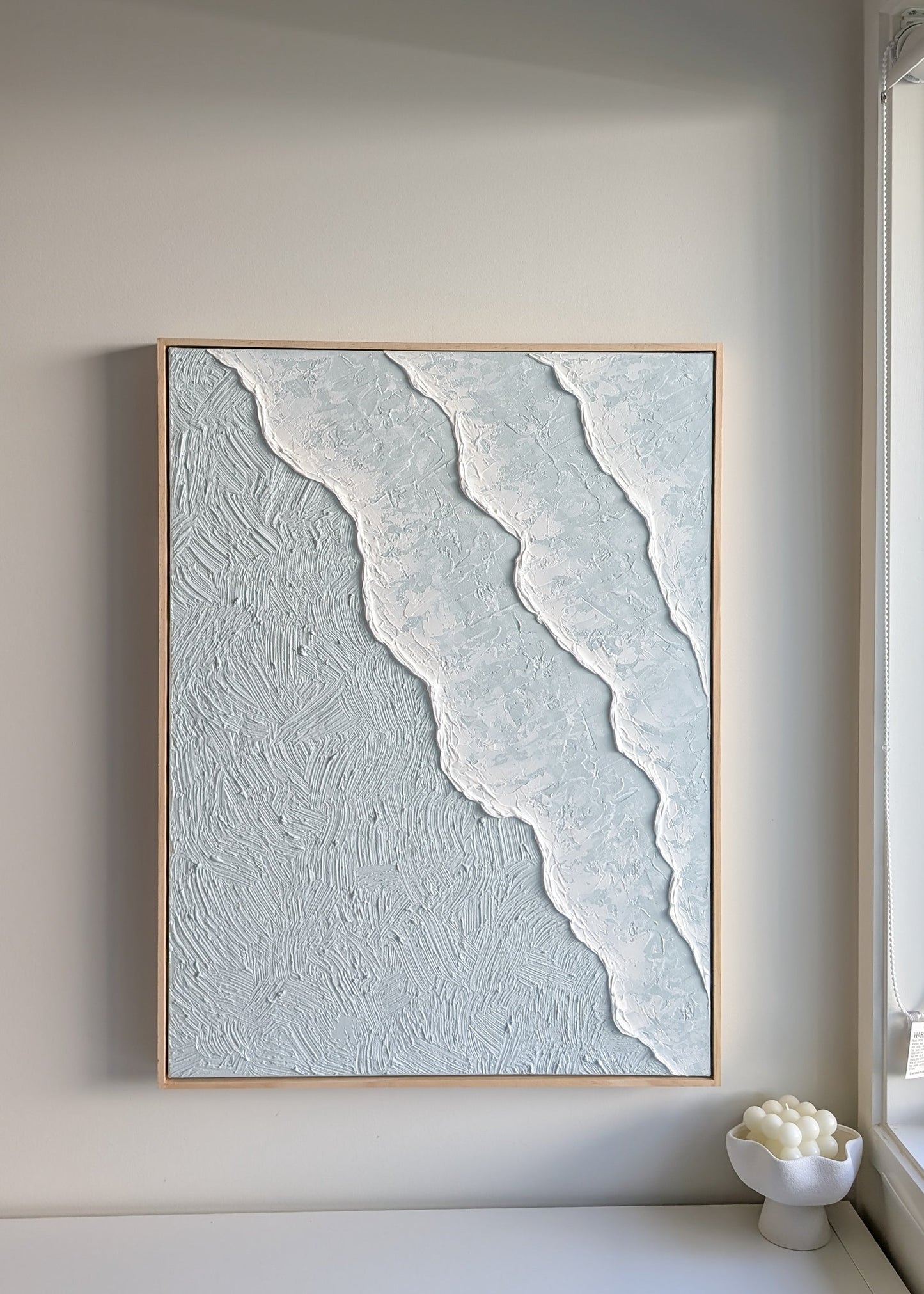Framed ocean waves textured pastel blue artwork on a wall in a minimalistic room