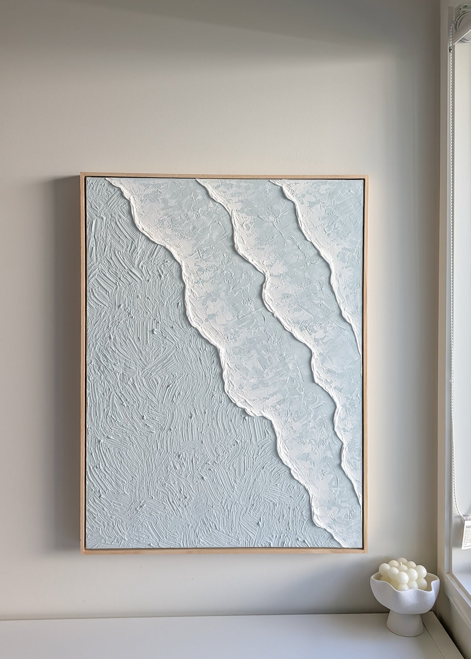 Framed ocean waves textured pastel blue artwork on a wall in a minimalistic room