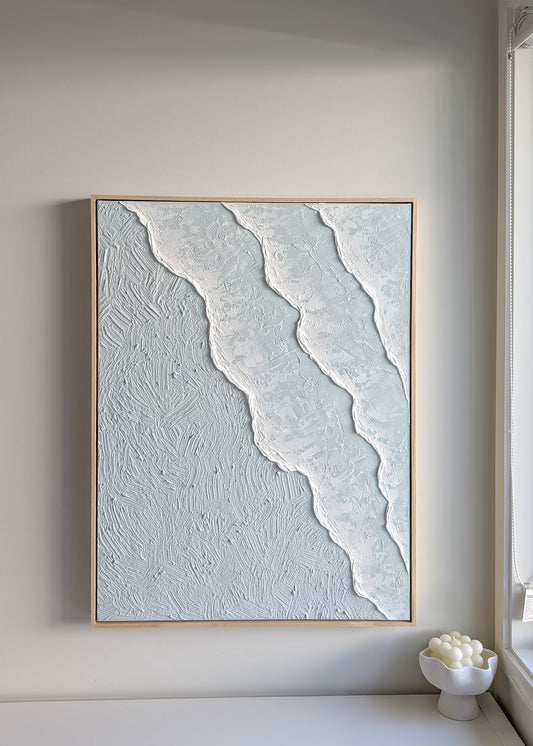 Framed ocean waves textured pastel blue artwork on a wall in a minimalistic room