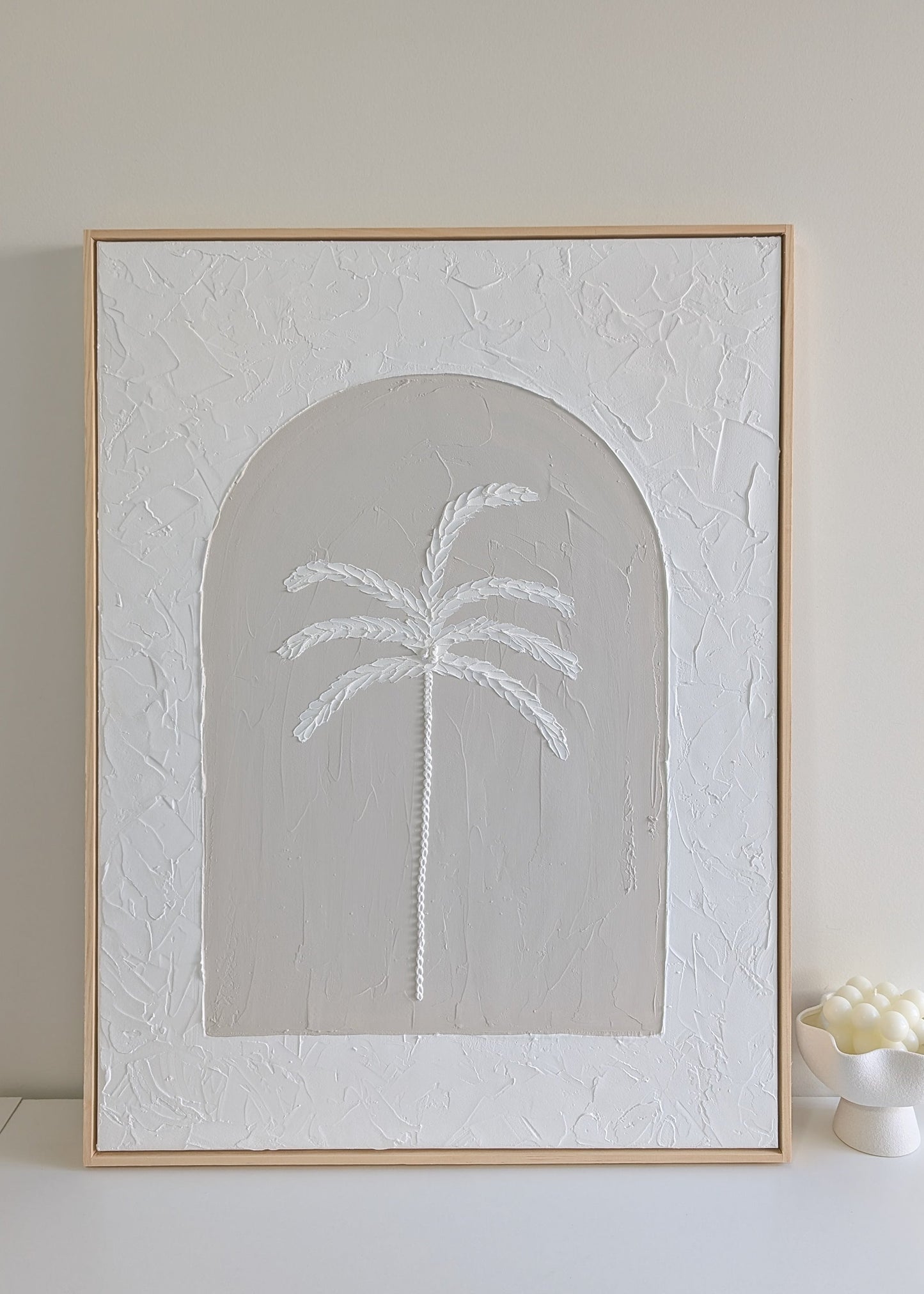Textured palm tree wall art on large neutral canvas with arched design and raw pinewood frame, displayed on a white shelf in natural light.