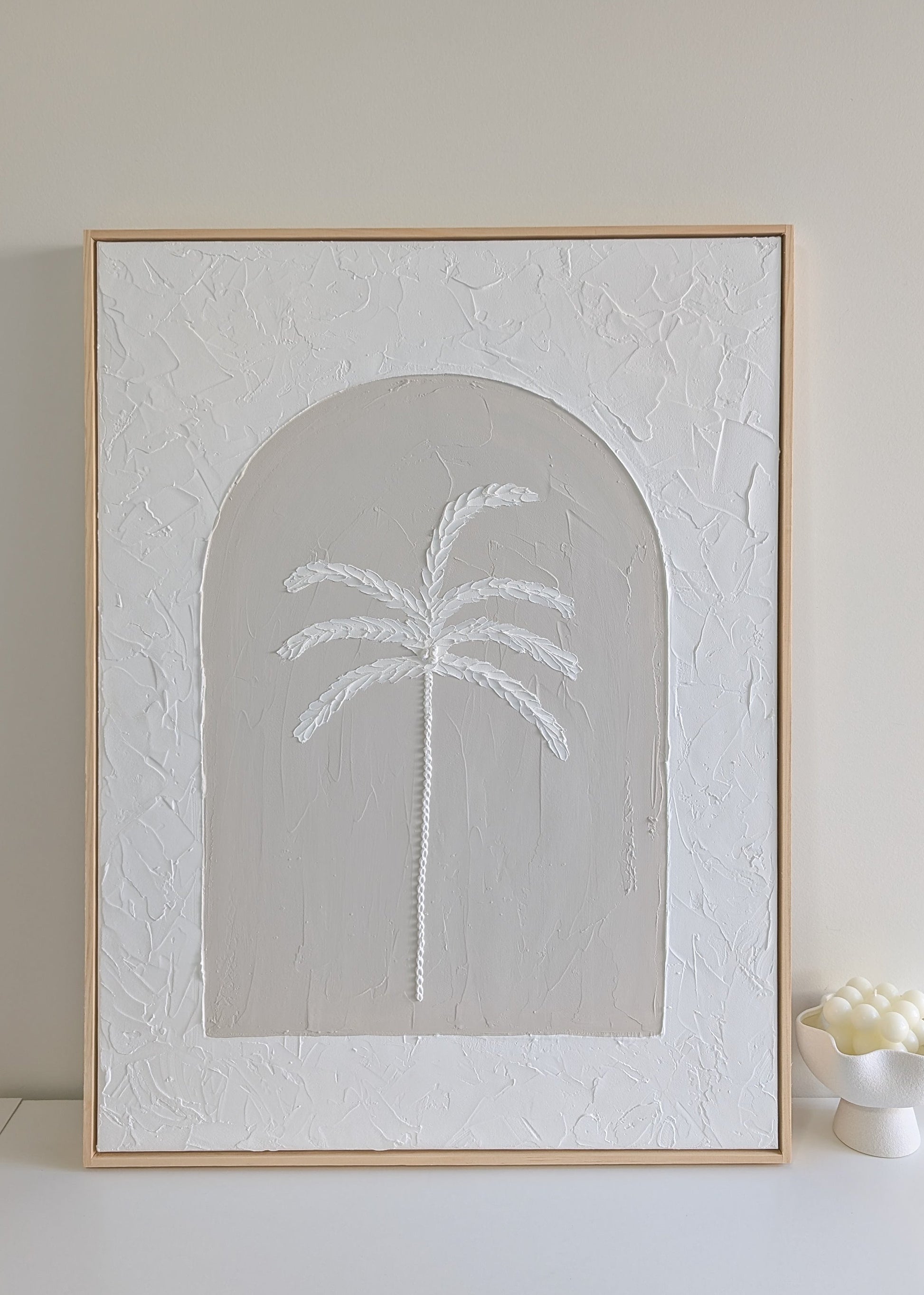 Textured palm tree wall art on large neutral canvas with arched design and raw pinewood frame, displayed on a white shelf in natural light.
