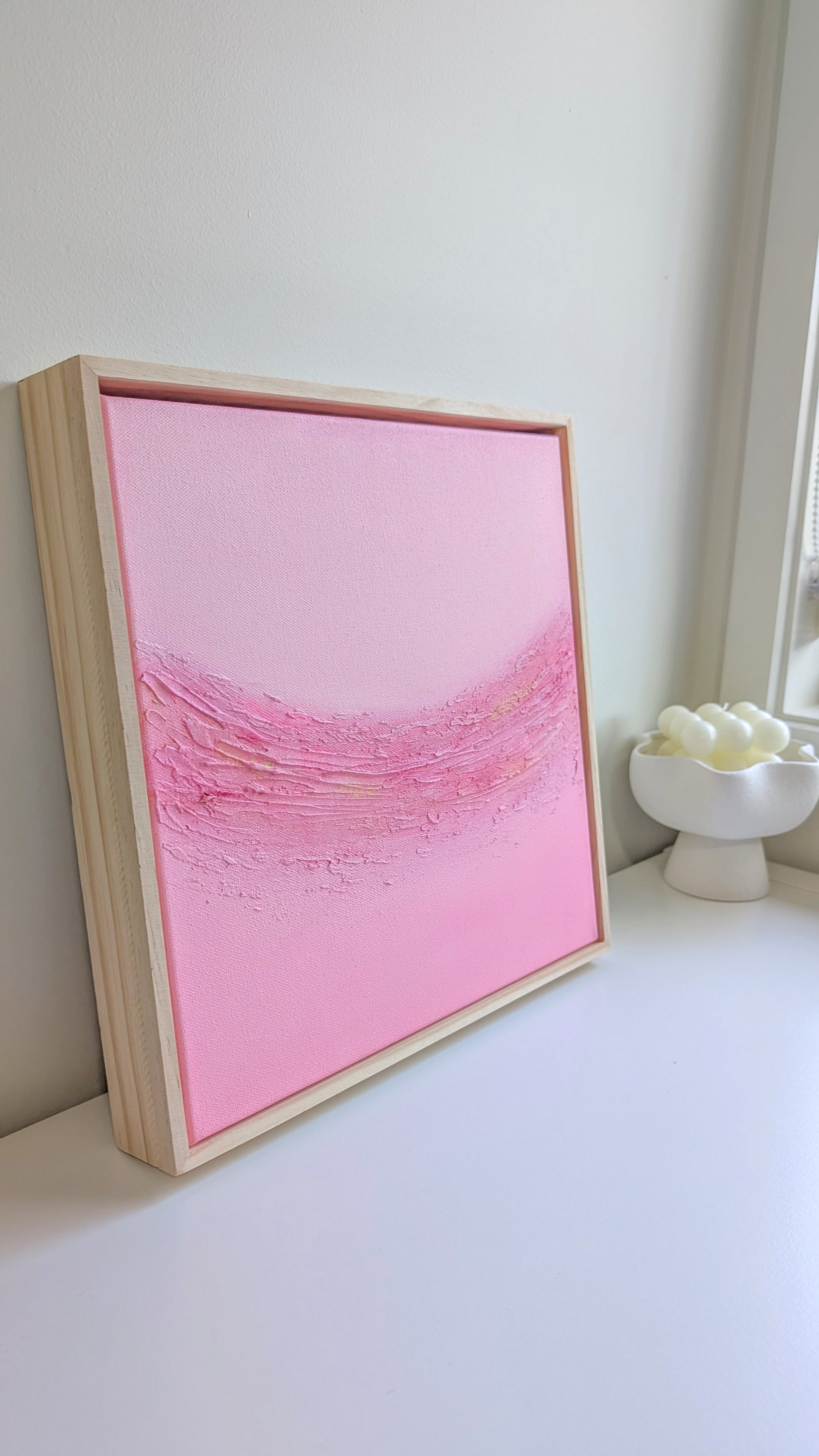 Close-up side view of "Blush Horizon" pink textured painting by Christen Rose, framed in pinewood and styled beside a ceramic flower decor.